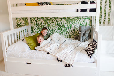 Two smiling boys in a white bunk bed with tropical leaf wallpaper, striped quilt, green pillows and a plush elephant in a modern kids bedroom