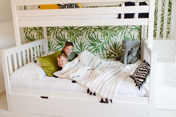 Two smiling boys in a white bunk bed with tropical leaf wallpaper, striped quilt, green pillows and a plush elephant in a modern kids bedroom