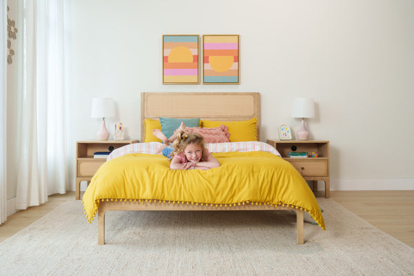 Smiling young girl lying on yellow comforter in bright modern kids' bedroom with wooden bedframe, bedside tables, lamps and abstract wall art