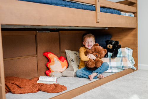 A young boy holding a teddy bear sitting under a low loft bed. Near him are various pillows, a blanket, another stuffed bear, and a book.