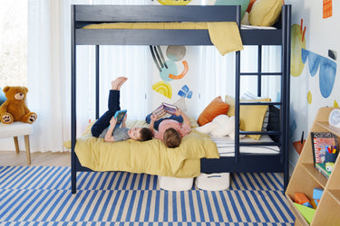 Two children lying on a lower bunk bed reading and using tablets in a bright modern kids’ bedroom with navy bunk, yellow bedding, playful wall art, and a blue-striped rug