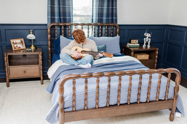 Red-haired teen playing a ukulele on a wooden spindle bed in a navy-blue bedroom with plaid curtains, nightstands and a toy robot