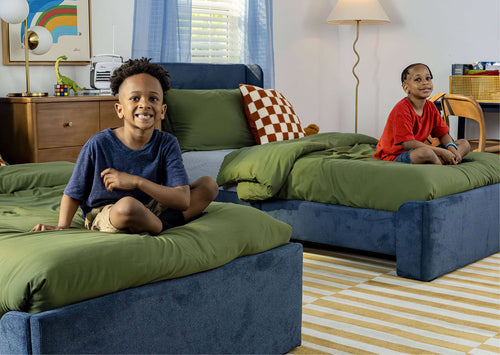 Two smiling boys on green beds in a bright kids' bedroom with blue curtains, checkered pillow, dinosaur toy and Rubik's Cube — children's room decor and boys' bedroom ideas.