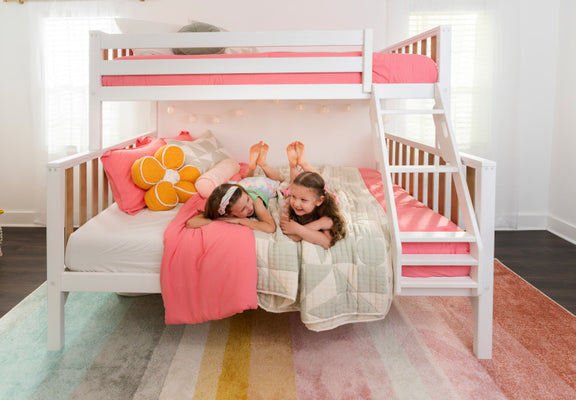 Two young girls lying on the lower bunk of a white wooden bunk bed with pink bedding, a flower-shaped throw pillow, stairs and a colorful striped rug in a bright kids' bedroom.