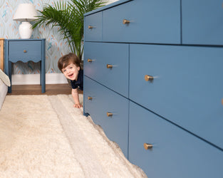 Toddler peeking behind a blue dresser in a modern kids' bedroom with bird-pattern wallpaper, blue nightstand, white lamp, indoor plant and plush rug
