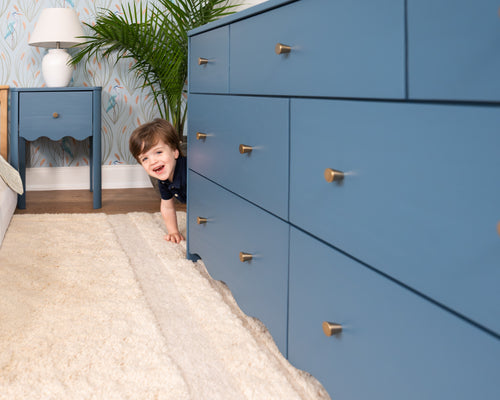 Toddler peeking behind a blue dresser in a modern kids' bedroom with bird-pattern wallpaper, blue nightstand, white lamp, indoor plant and plush rug