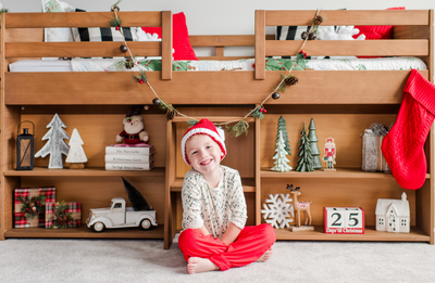 Child in Christmas pajamas and Santa hat sitting in front of wooden bunk bed decorated with festive ornaments, garland, Christmas stocking, and holiday-themed bookshelf decorations.