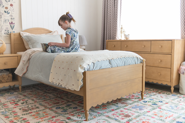 Girl reading on a wooden bed in a cozy children's bedroom with striped bedding, floral accents, wooden dresser, patterned rug, and soft natural light, ideal kids bedroom decor