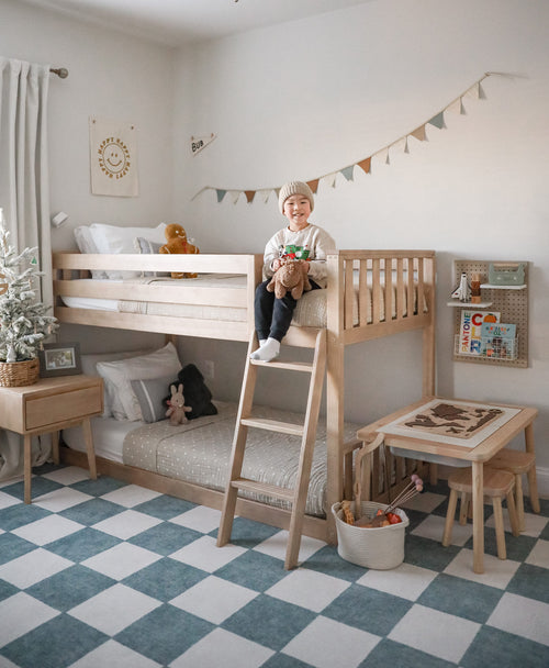 Young child sitting on a natural wood bunk bed in a cozy neutral Scandinavian-style kids' bedroom with a blue-and-white checkered rug, play table, stuffed animals and bunting.