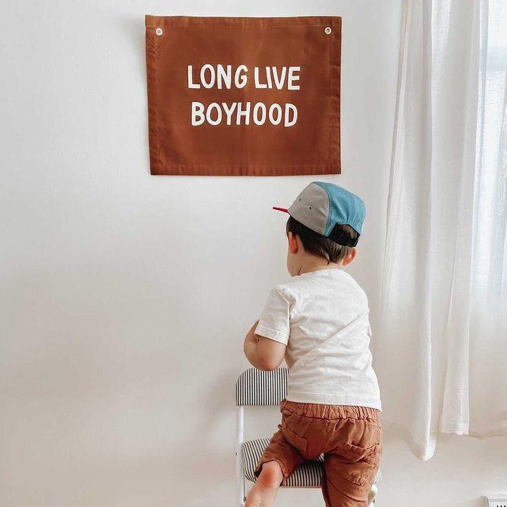 Young boy in a cap looking at a "Long Live Boyhood" banner in a bright room.