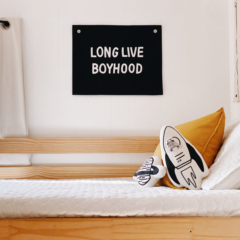 Cozy boys' bedroom with a wooden bed, astronaut-themed pillows, and a "Long Live Boyhood" wall banner.