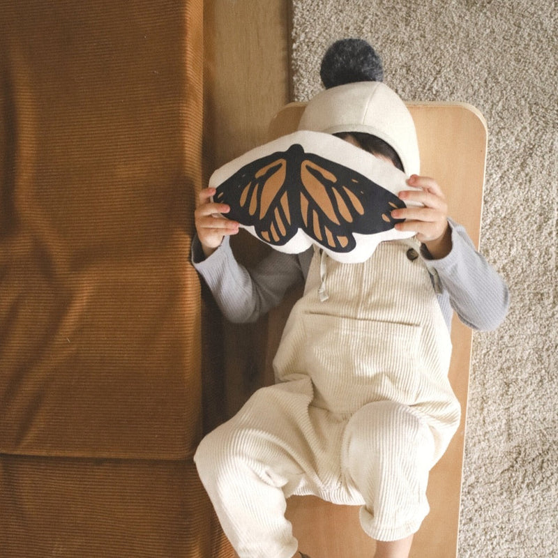 Child in cozy outfit laying down, holding a butterfly-shaped pillow, playful and cute scene.