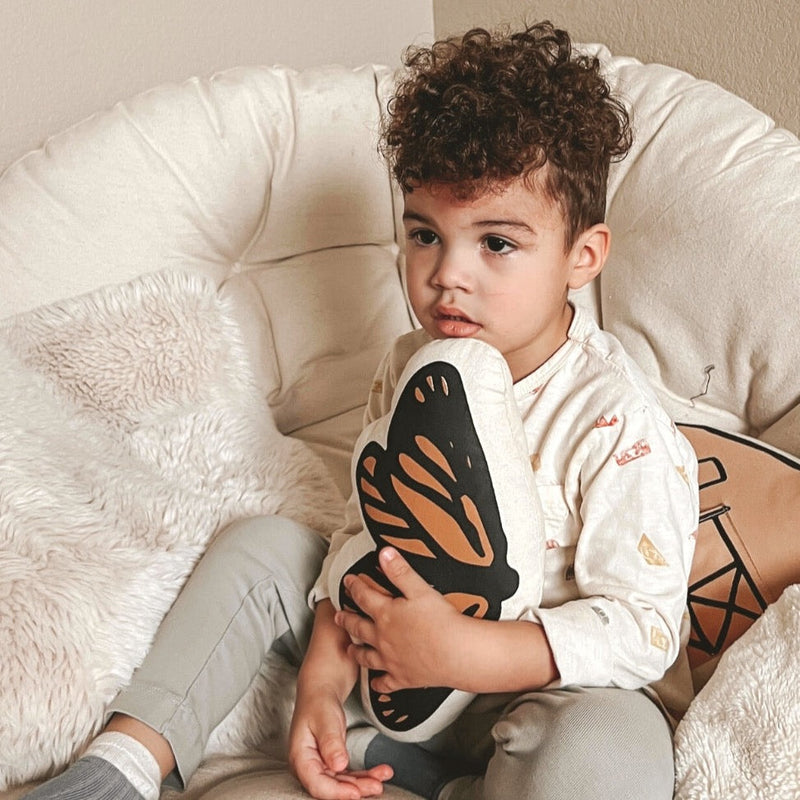 Toddler with curly hair holding a butterfly pillow on a cozy chair.