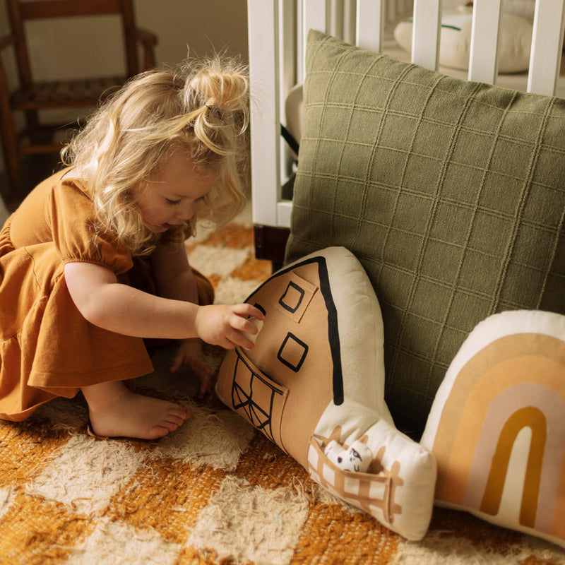 Toddler playing with house-shaped cushion in cozy nursery setting.