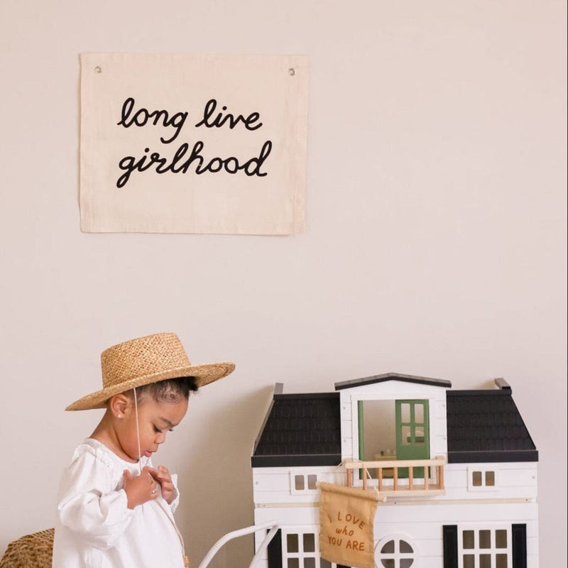 Young child wearing a straw hat playing with toy dollhouse under "long live girlhood" banner.
