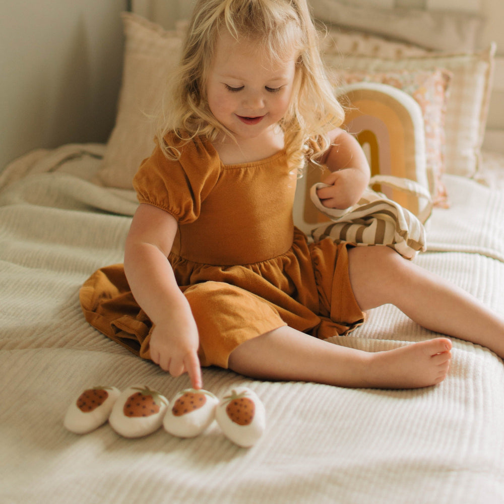 Young child in a mustard dress playing with strawberry-shaped toys on a bed.
