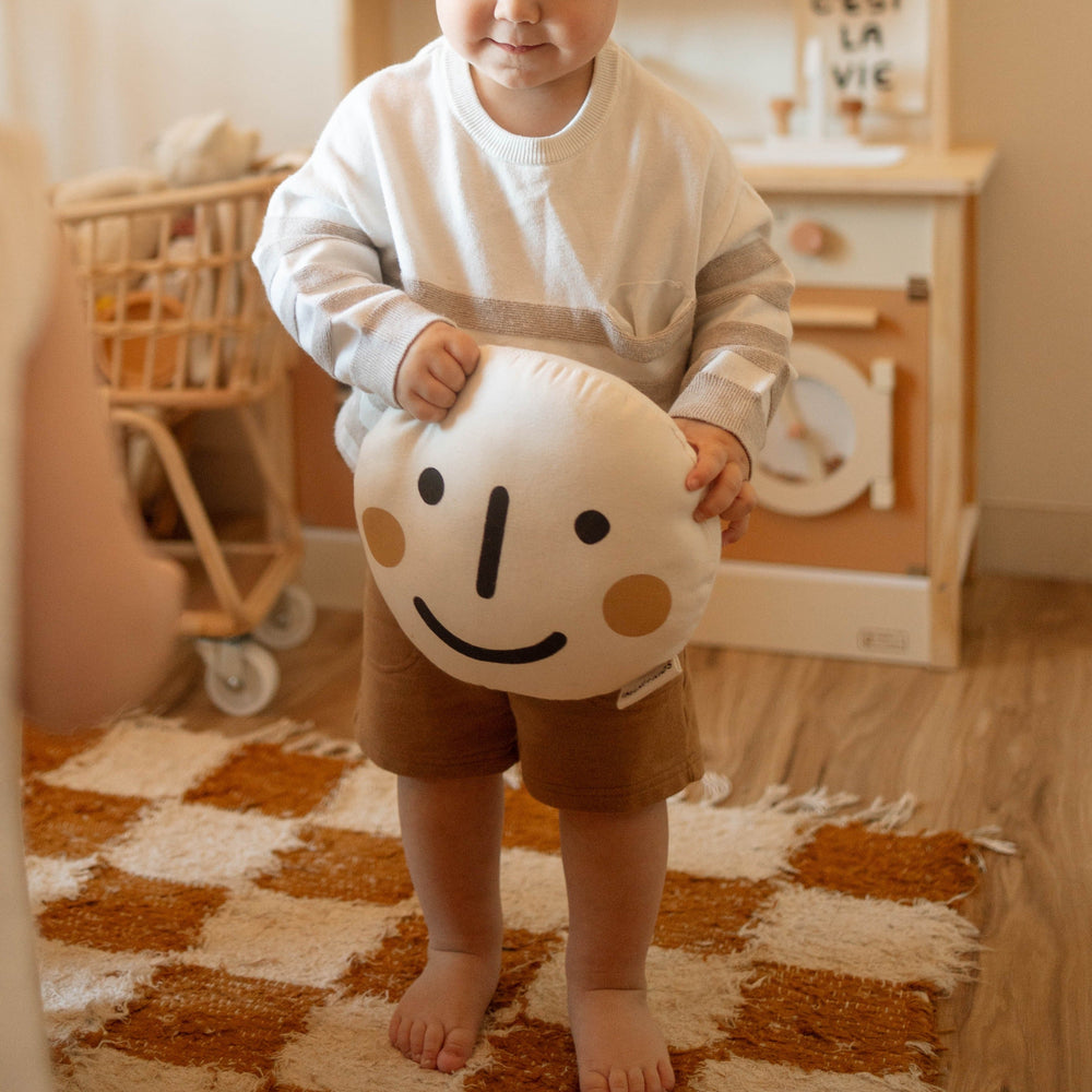 Child holding a smiley face cushion in a cozy playroom.