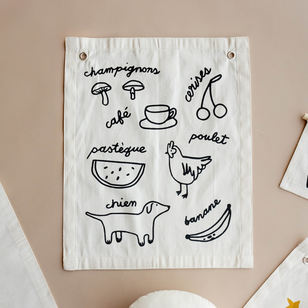 Fabric wall hanging with French food illustrations and text including mushrooms, cherries, coffee, watermelon, chicken, dog, and banana.