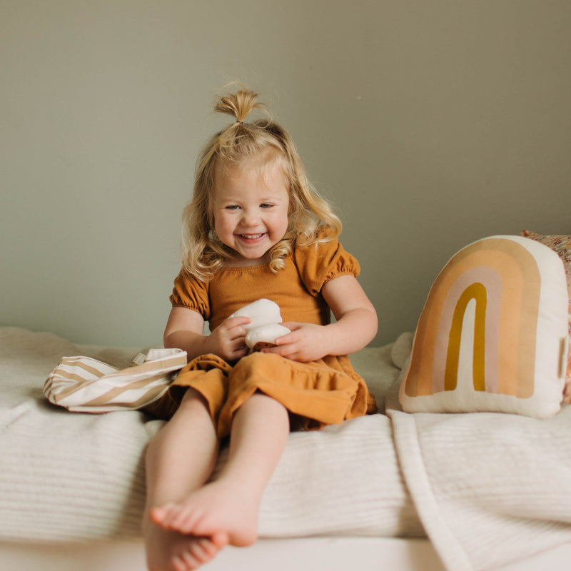 Smiling toddler in a mustard dress sitting on a bed with a rainbow pillow.