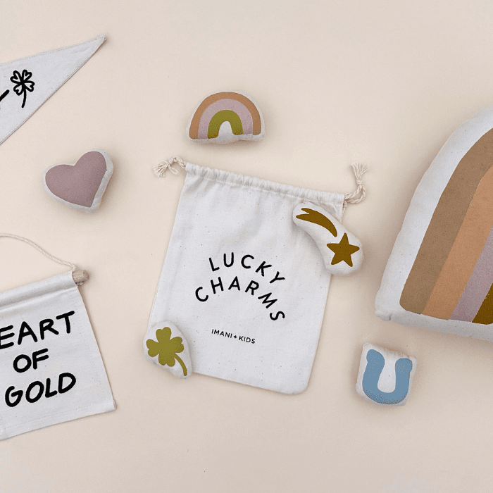 Cloth drawstring bag labeled "Lucky Charms" surrounded by pastel-shaped plush toys including a rainbow, heart, and horseshoe on a beige background.