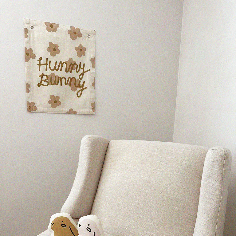 Cozy nursery corner with beige chair, decorative "Hunny Bunny" wall hanging, plush toys, and floral pattern.