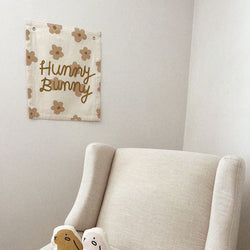 Cozy nursery corner with beige chair, decorative "Hunny Bunny" wall hanging, plush toys, and floral pattern.
