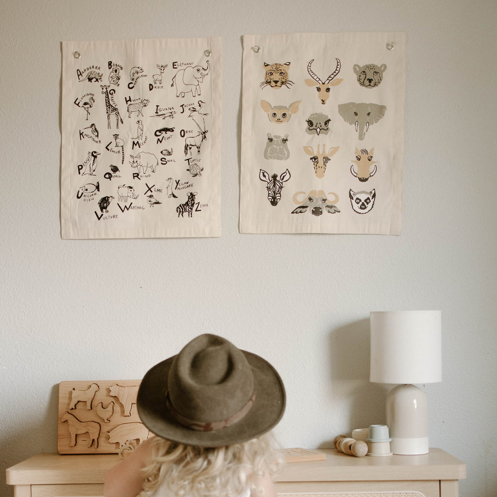 Child in hat looking at animal alphabet posters on wall above wooden toy and lamp.