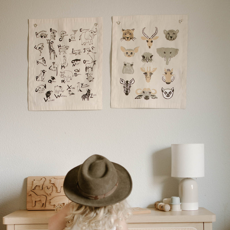 Child in hat looking at animal alphabet posters on wall above wooden toy and lamp.