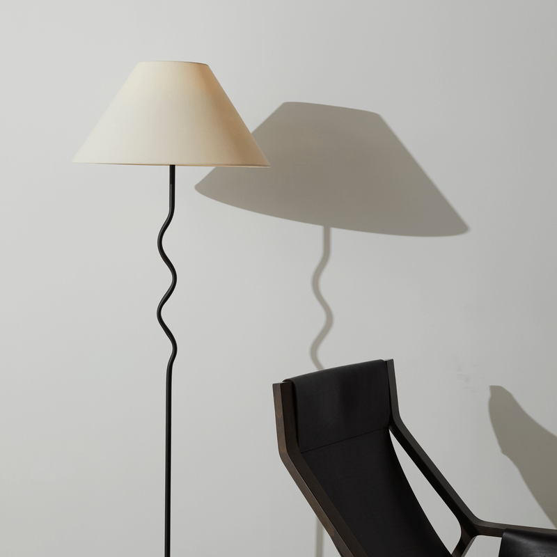 Modern floor lamp with cream shade beside a black minimalist chair against a neutral background.