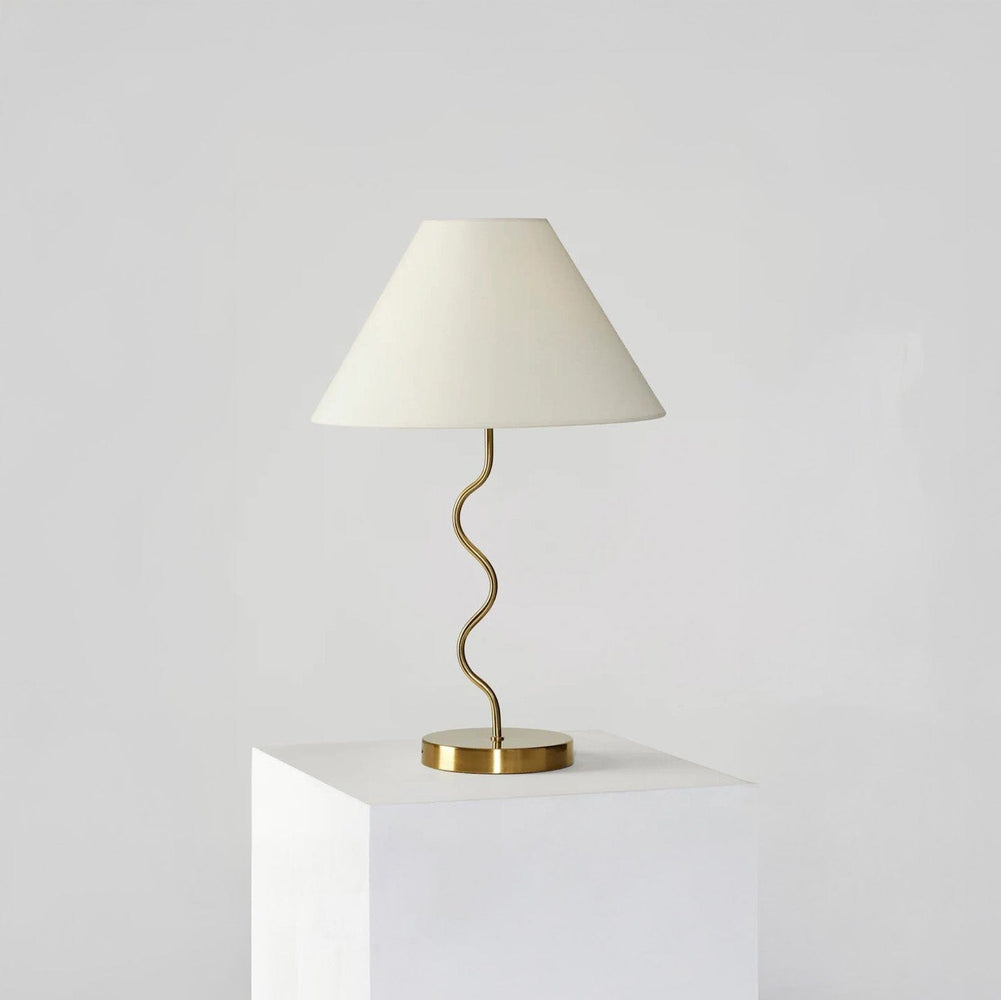 Stylish modern table lamp with a wavy gold stand and white lampshade on a minimalist pedestal, perfect for contemporary home decor.