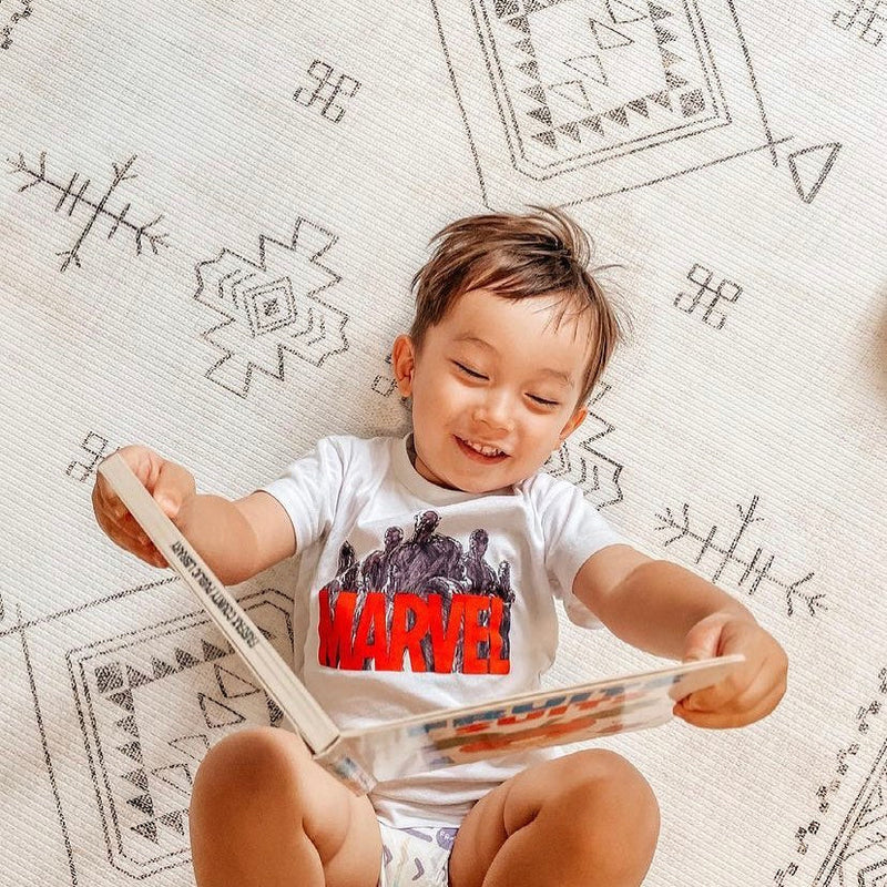 Toddler reading a Marvel comic book while smiling on patterned carpet.