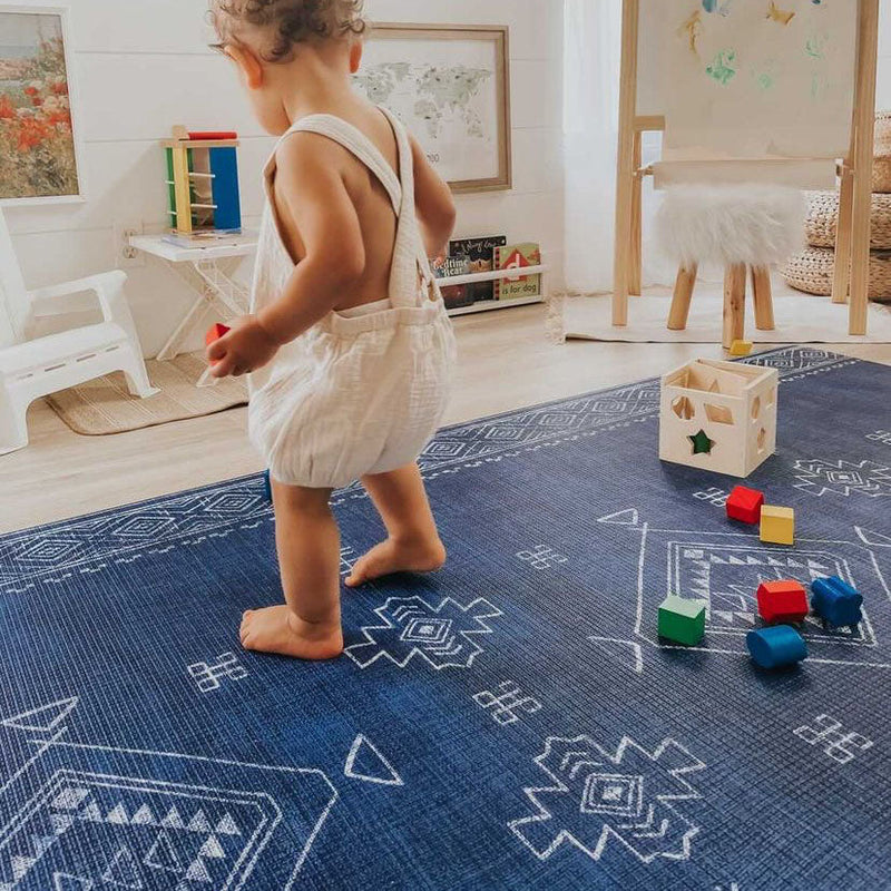 Toddler playing on a patterned blue rug with colorful blocks in a nursery room.