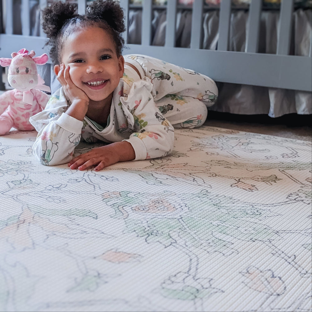 Toddler in floral pajamas smiling on a patterned play mat with a pink giraffe stuffed animal nearby in a nursery.
