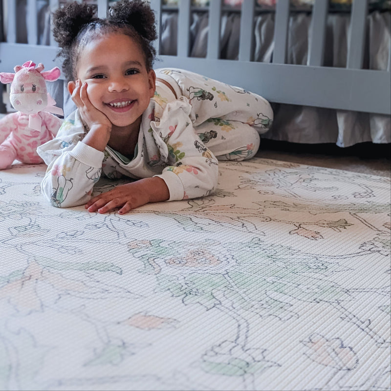 Toddler in floral pajamas smiling on a patterned play mat with a pink giraffe stuffed animal nearby in a nursery.