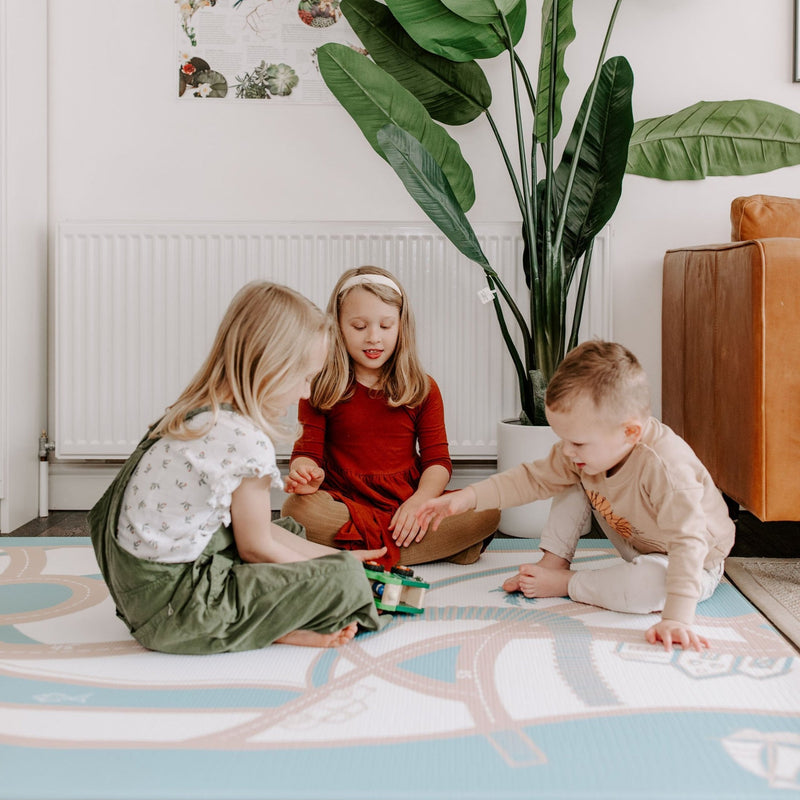 Children playing together on a colorful mat in a bright living room with a large potted plant.