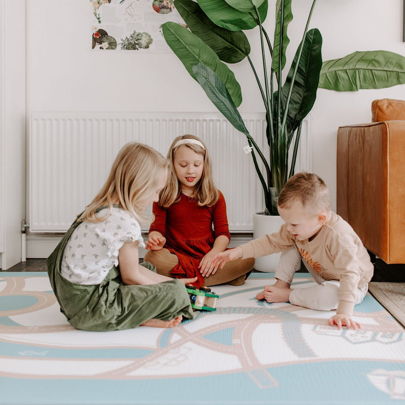 Children playing on a colorful mat in a cozy living room with a large plant nearby.