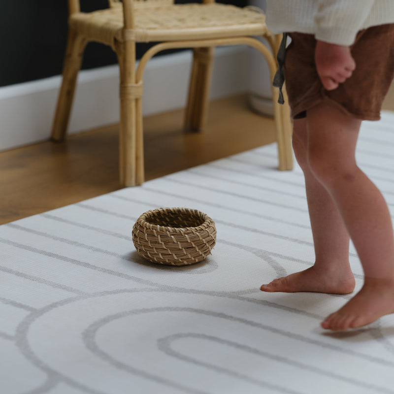 Child standing on a patterned rug next to a wicker basket and wooden chair.