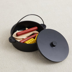 Toy cooking set in black pot with lid, featuring wooden vegetables and sausages, on beige carpet.