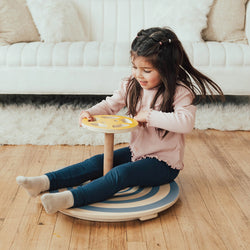 Young girl playing with a spinning balance toy indoors on a wooden floor, promoting children's sensory and motor skills development.
