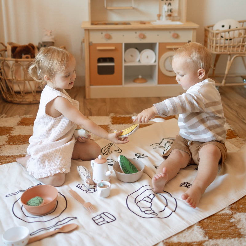 Two toddlers playing in a cozy playroom with a toy kitchen and picnic setup.