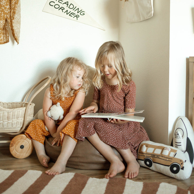 Two young children reading together in a cozy playroom setting with toys and cushions.