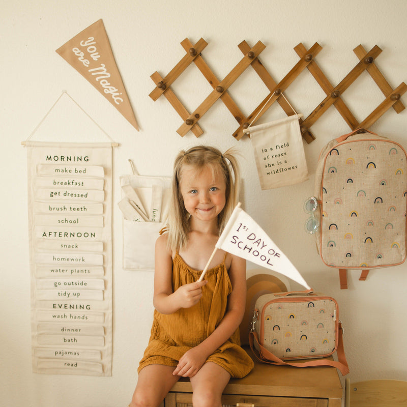 Girl smiling and holding a "1st Day of School" flag in cozy, decorated room with wall hangings and backpacks.