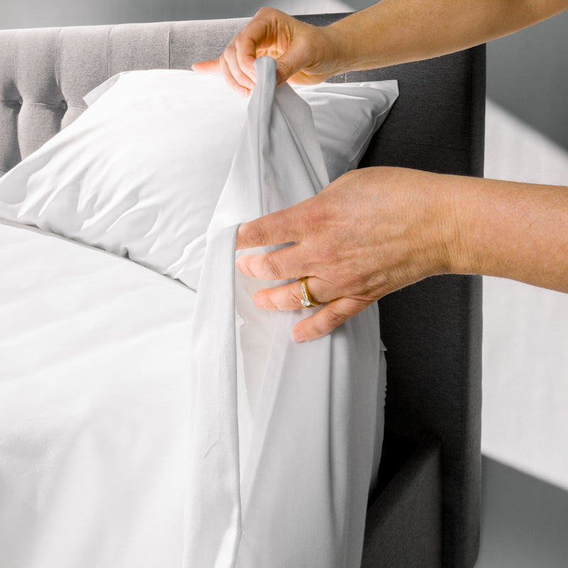 Hands adjusting a white fitted sheet on a bed with a grey upholstered headboard.