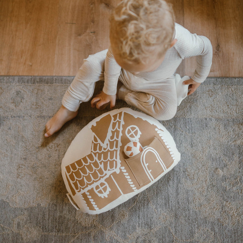 Toddler playing with gingerbread house pillow on rug in cozy home setting.