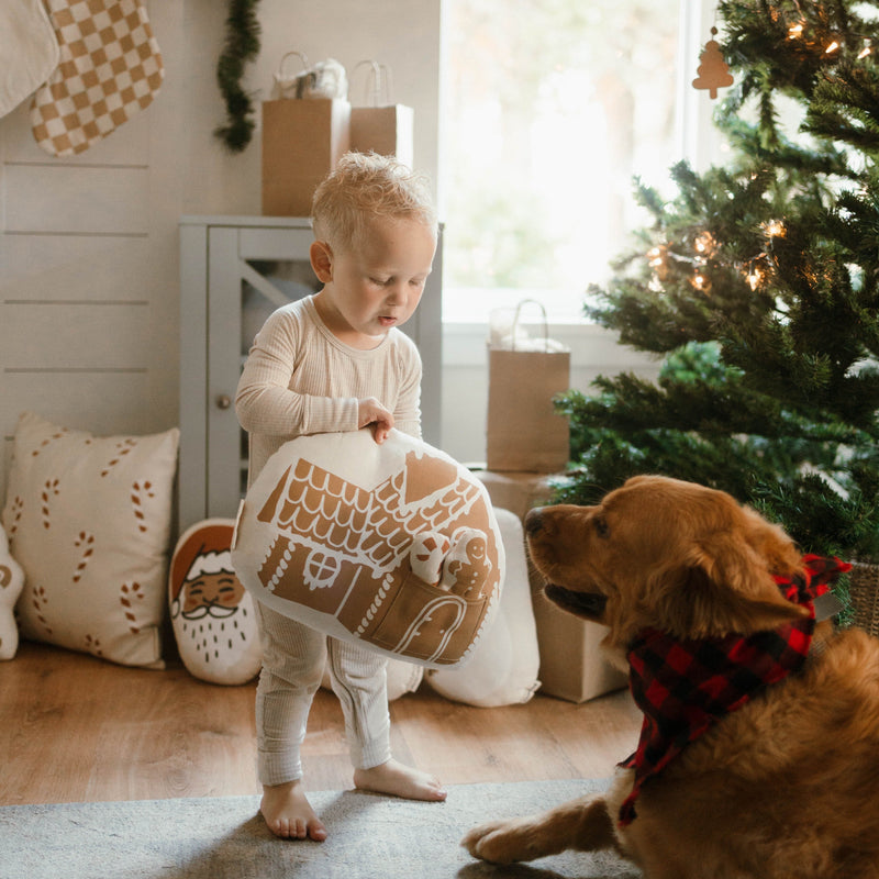 Young child holding a gingerbread-themed pillow near a Christmas tree with a golden retriever wearing a red plaid scarf.