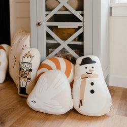 Plush holiday-themed pillows featuring a snowman, bird, nutcracker, and candy cane in a cozy living room setting.