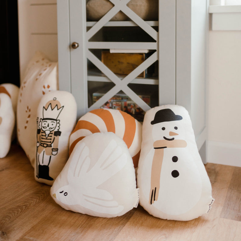 Plush holiday-themed pillows featuring a snowman, bird, nutcracker, and candy cane in a cozy living room setting.