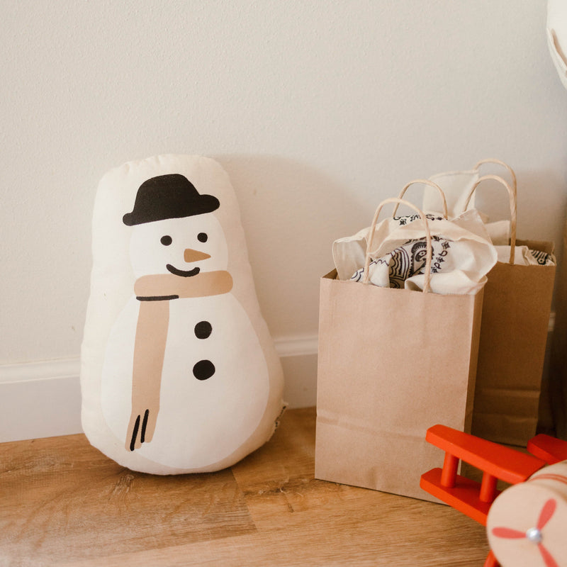 Stuffed snowman toy next to brown paper gift bags and wooden toy airplane on wooden floor.