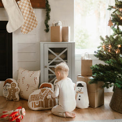 Toddler enjoying Christmas decorations beside a holiday-themed pillow and Christmas tree in a cozy living room.