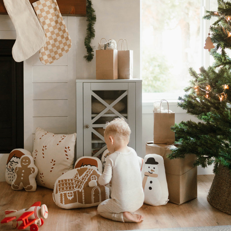 Toddler enjoying Christmas decorations beside a holiday-themed pillow and Christmas tree in a cozy living room.